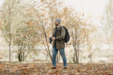 Aynalı gözlüklü, güneş gözlüklü, zeytin askeri ceketli, kol saati olan sırt çantalı cesur bir gezgin DSR kamerasını tutuyor ve vahşi ormana bakıyor.