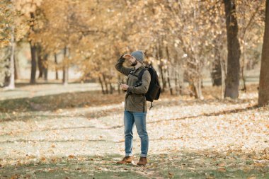 Zeytin askeri kargo savaş ceketli, kot pantolonlu, sırt çantası ve kol saati olan bir gezgin öğle vakti DSR kamerasını ve akıllı telefonu elinde tutuyor. Bir fotoğrafçı birini arıyor..