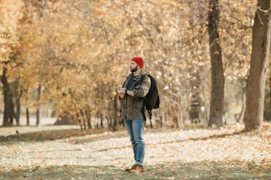 A photographer with a beard in an olive military combat cargo jacket, jeans, red hat with backpack holds the DSLR camera and his wristwatch and looks away in the forest at the afternoon