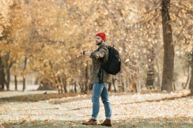 Zeytin askeri kargo ceketli, kot pantolonlu, sırt çantalı kırmızı şapkalı bir fotoğrafçı öğleden sonra ormandaki kol saatinin saatini kontrol ediyor.