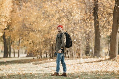 Zeytin askeri kargo ceketli, kot pantolonlu, sırt çantalı ve kol saatli kırmızı şapkalı bir foto muhabiri profesyonel DSR kamerasını tutuyor ve öğle vakti ormanda bekliyor.
