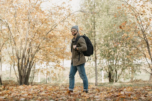 A brutal traveler with a beard in aviator sunglasses with mirror lenses, olive military combat jacket, jeans, hat with backpack and wristwatch walks in the afternoon in the forest. A photojournalist.
