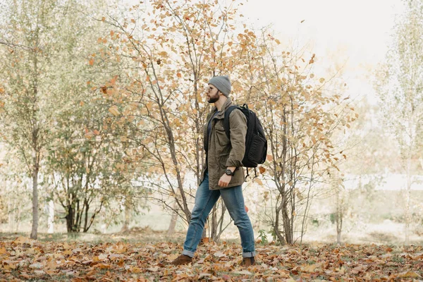 A brutal traveler with a beard in aviator sunglasses with mirror lenses, olive military combat jacket, jeans, hat with backpack and wristwatch goes through the forest at the noon. A photojournalist.