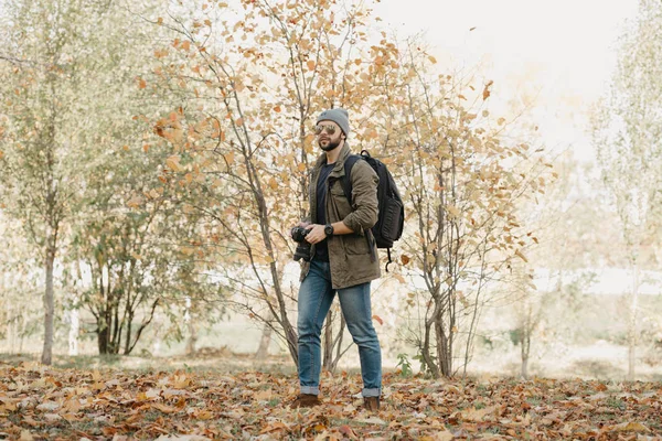 A photojournalist with a beard in aviator sunglasses with mirror lenses, olive military jacket, jeans, hat with a backpack with wristwatch holds the DSLR camera, looks around near the battlefield