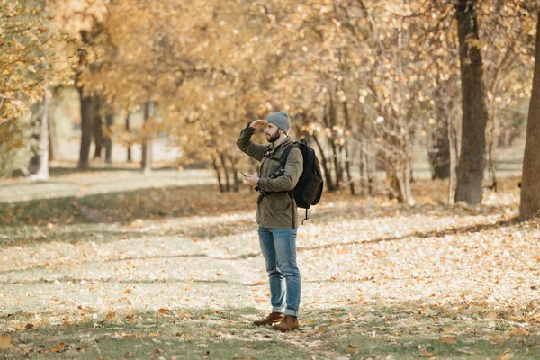 A traveler with a beard in an olive military cargo combat jacket, jeans, hat with backpack and wristwatch holds the DSLR camera and the smartphone. A Photographer looks straight  at the noon