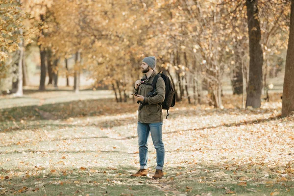 A traveler with a beard in an olive military cargo combat jacket, jeans, hat with backpack and wristwatch holds the DSLR camera and the smartphone. A Photographer waits for someone at noon.