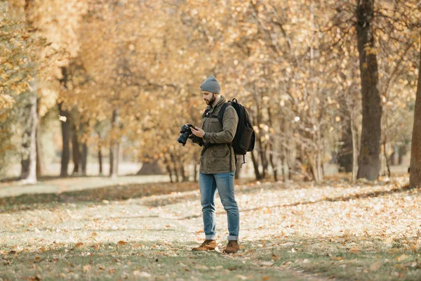 A traveler with a beard in an olive military cargo combat jacket, jeans, hat with backpack and wristwatch holds the DSLR camera and checks something in the smartphone in the forest at the noon.