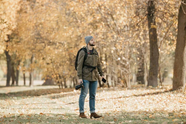 A photographer with a beard in an olive military cargo combat jacket, jeans, hat with backpack and wristwatch holds the DSLR camera, the smartphone and waits in the forest at the noon.