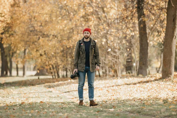 A photographer with a beard in an olive military combat jacket, jeans, red hat with backpack and wristwatch holds the professional DSLR camera and waits in the forest at the afternoon