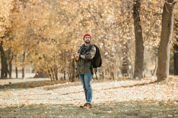 A brave photographer with a beard in an olive military combat jacket, jeans, red hat with backpack holds the DSLR camera and his wristwatch in the forest at the afternoon