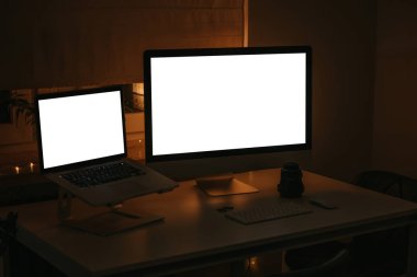 Masaüstü bilgisayarı, masadaki dizüstü bilgisayar, iki hafıza kartı, geniş açılı lens, klavye ve fare ışığın kapalı olduğu odada pencerenin yanındaki beyaz masada duruyor. 