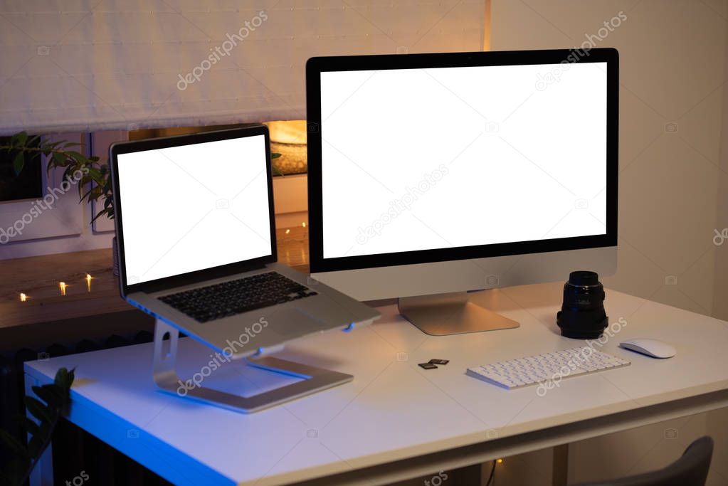 Modern workspace. All-in-one computer, laptop on the stand, 2 memory cards, wide-angle lens, keyboard, mouse stands on the white table near the window with lamp and garland. Blue light from the left.