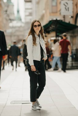 Beyaz kot ceketli, siyah kotlu, gri spor ayakkabılı ve güneş gözlüklü güzel bir kadın blogcu tripodu antik Avrupa sokaklarının ortasında tutuyor.