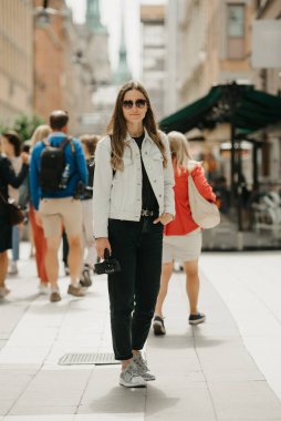 Beyaz kot ceketli, siyah kotlu, gri spor ayakkabılı ve güneş gözlüklü muhteşem bir kadın blogcu tripodu antik Avrupa sokaklarının ortasında tutuyor.