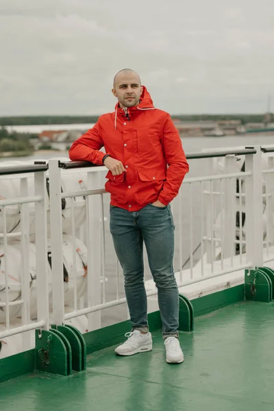 Bald brutal man with stubble in a red jacket, blue jeans and white sneakers on the ship enjoying his cruise in the cloudy afternoon