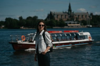 Beyaz kot ceketli, siyah kotlu, gri spor ayakkabılı, güneş gözlüklü ve sırt çantalı genç bir bayan turist, Avrupa 'nın göl kıyısındaki merkezinde geminin arka planında poz veriyor.