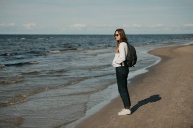 Beyaz kot ceket, siyah kot pantolon, gri spor ayakkabı, güneş gözlüğü ve sırt çantasıyla gezen genç bir bayan turist