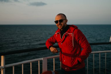 Güneş gözlüklü kırmızı ceketli kel, mutlu adam gün batımını seyreden bir gemide poz veriyor.