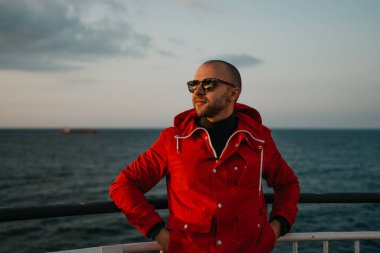 Kapüşonlu, kırmızı ceketli ve güneş gözlüklü şık bir adam gemide günbatımının tadını çıkarıyor. Arka planda kuru kargo gemisi var..