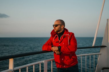 Güneş gözlüklü, kirli sakallı, kel bir adam. Elleri katlanmış, gün batımını seyrediyor..