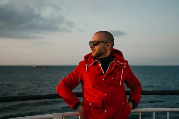 A bald man with stubble in a red jacket in the sunglasses with hands in pockets aboard a ship looking at the sunset. Dry cargo ship in the background.