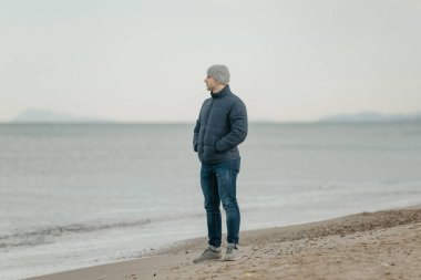 Gri şapkalı, kotlu, salam ceketli ve spor ayakkabılı kaslı bir adam Akdeniz 'e bakıyor.