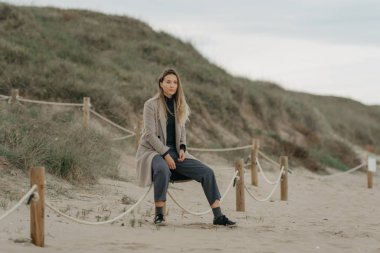 Uzun saçlı, ceketli, pantolonlu ve spor ayakkabılı güzel bir kadın sahilde otururken poz veriyor. Arka planda kum tepeleri.