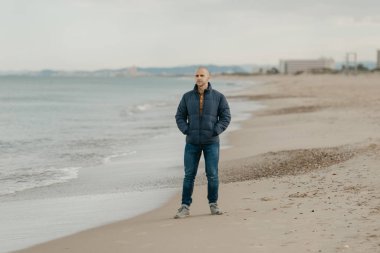 Kot pantolon, salam ceketi ve spor ayakkabısı giymiş kaslı kel bir adam Akdeniz kıyısında elleri ceplerinde bekliyor.