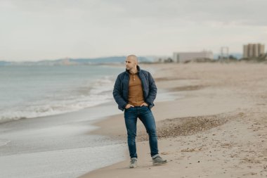 Bologna ceketli, spor ayakkabılı, kaslı, kel bir adam Akdeniz kıyısında kot pantolon cebinde bekliyor.