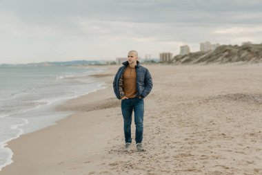 Kas yığını, kel, ceketli, kot pantolonlu ve spor ayakkabılı bir adam Akdeniz 'e yakın kot pantolonlarının cebinde düz duruyor.