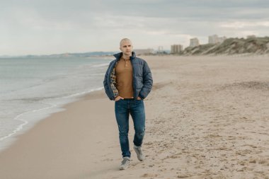 Kas yığını, kel, ceketli, kot pantolonlu ve spor ayakkabılı bir adam Akdeniz kıyısında öğleden sonra kot pantolon cebinde yürür. Arka planda kum tepeleri.