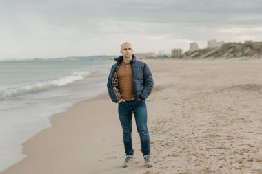 Aşağı ceketli, kot pantolonlu ve spor ayakkabılı, kaslı, kel bir adam Akdeniz kıyısında öğleden sonra kot pantolon cebinde yürür. Arka planda kum tepeleri.