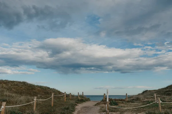 A walkway between sandy green dunes to the Mediterranean Sea in the afternoon. Clouds on the sky.
