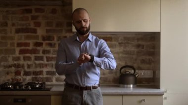 A bald brutal businessman with a beard in a blue shirt, grey trousers looks on his wristwatch, then puts his hands in his pockets in the kitchen