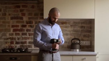 A bald photographer searches for some preferences in his camera then looks straight at the camera with a smile. A man with a beard wears two silver earrings, a blue shirt, grey trousers, and a brown leather belt.