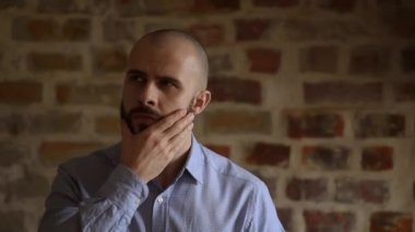 A close portrait of a serious businessman who thinks about something and strokes his beard. A man wears two silver earrings and a blue shirt.