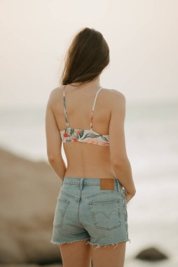 Uzun saçlı, güzel bir manken. Sırtında elleri ile kızıl denizin kayalık sahilindeki kot şortların ceplerine poz veriyor. Güneşin doğuşunda yüzer vaziyette.