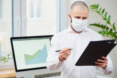 Coronavirus 'a karşı tıbbi maske takan bir adam (COVID-19). Çalışma alanında elinde kalem ve dosya tutan bir ofis çalışanı. Arka planda bilgisayarlar ve yeşil bitkiler var. Coronavirus karantinası