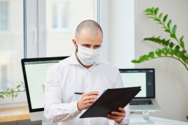 Coronavirus 'a karşı tıbbi maske takan bir adam (COVID-19). Bir ofis çalışanı çalışma alanında bilgisayarlarla ve arka planda yeşil bitkilerle notlar alıyor. Coronavirus karantinası