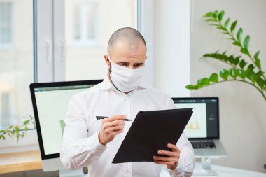 Coronavirus 'a karşı tıbbi maske takan bir adam (COVID-19). Arka planda bilgisayarlar ve yeşil bitkilerle çalışma alanında notlar alan bir yönetici. Coronavirus karantinası.