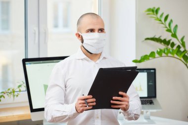 Coronavirus 'a karşı tıbbi maske takan bir adam (COVID-19). Çalışma alanında elinde kalem ve klasör olan bir mühendis. Arkasında bilgisayarlar ve yeşil bitkiler var. Coronavirus karantinası.