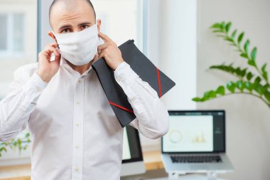 Koronavirüse karşı tıbbi prosedür maskesi çıkaran bir adam. Çalışma alanında elinde bilgisayarları ve arka planda yeşil bitkileri olan bir dosya tutan bir ofis çalışanı. COVID-19 karantinası.