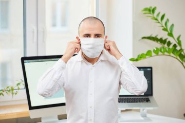 Beyaz gömlekli bir adam Coronavirus 'a (COVID-19) karşı tıbbi yüz maskesi çıkarıyor. Çalışma alanında bilgisayarları ve arka planda yeşil bitkileri olan bir ofis çalışanı. Coronavirus karantinası.