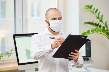 Coronavirus 'a karşı tıbbi maske takan bir adam (COVID-19). Kalem ve klasörlü bir mühendis çalışma alanında bilgisayarlarla ve arka planda yeşil bitkilerle notlar tutuyor. Coronavirus karantinası.