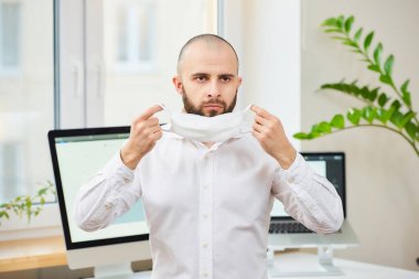Gömlekli sakallı bir adam, Coronavirus 'a karşı tıbbi maske takıyor. Çalışma alanında bilgisayarları ve arka planda yeşil bitkileri olan bir yönetici. Coronavirus karantinası.
