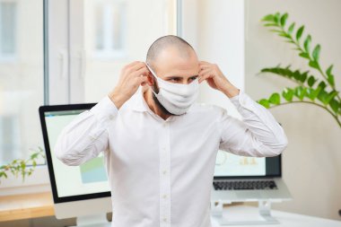 Gömlekli sakallı bir adam, Coronavirus 'a karşı tıbbi maske takıyor. İş yerinde bilgisayarları ve yeşil bitkileri olan bir mühendis. Coronavirus karantinası.