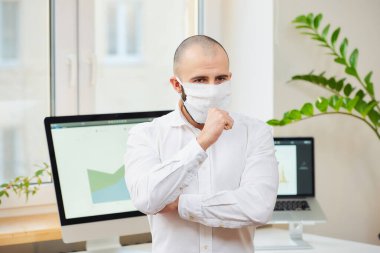  Tıbbi yüz maskesi takmış bir adam Coronavirus 'a karşı (COVID-19) yumruğuna öksürüyor. Çalışma alanında bilgisayarları ve arka planda yeşil bitkileri olan bir yönetici. Coronavirus karantinası.