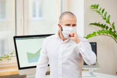 Tıbbi yüz maskesi takmış bir adam Coronavirus 'a karşı (COVID-19) yumruğuna öksürüyor. Çalışma alanında bilgisayarları ve arka planda yeşil bitkileri olan bir ofis çalışanı. Coronavirus karantinası.