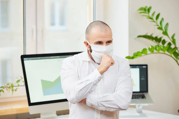 A man in a medical face mask against the coronavirus (COVID-19) strongly coughing into his fist. A manager at his workspace with computers and green plants in the background. Coronavirus quarantine.