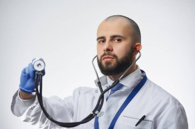 Elinde steteskop tutan ciddi bir doktorun portresi. Tek kullanımlık tıbbi eldiven. Beyaz önlüklü ve mavi kravatlı sakallı bir doktor..